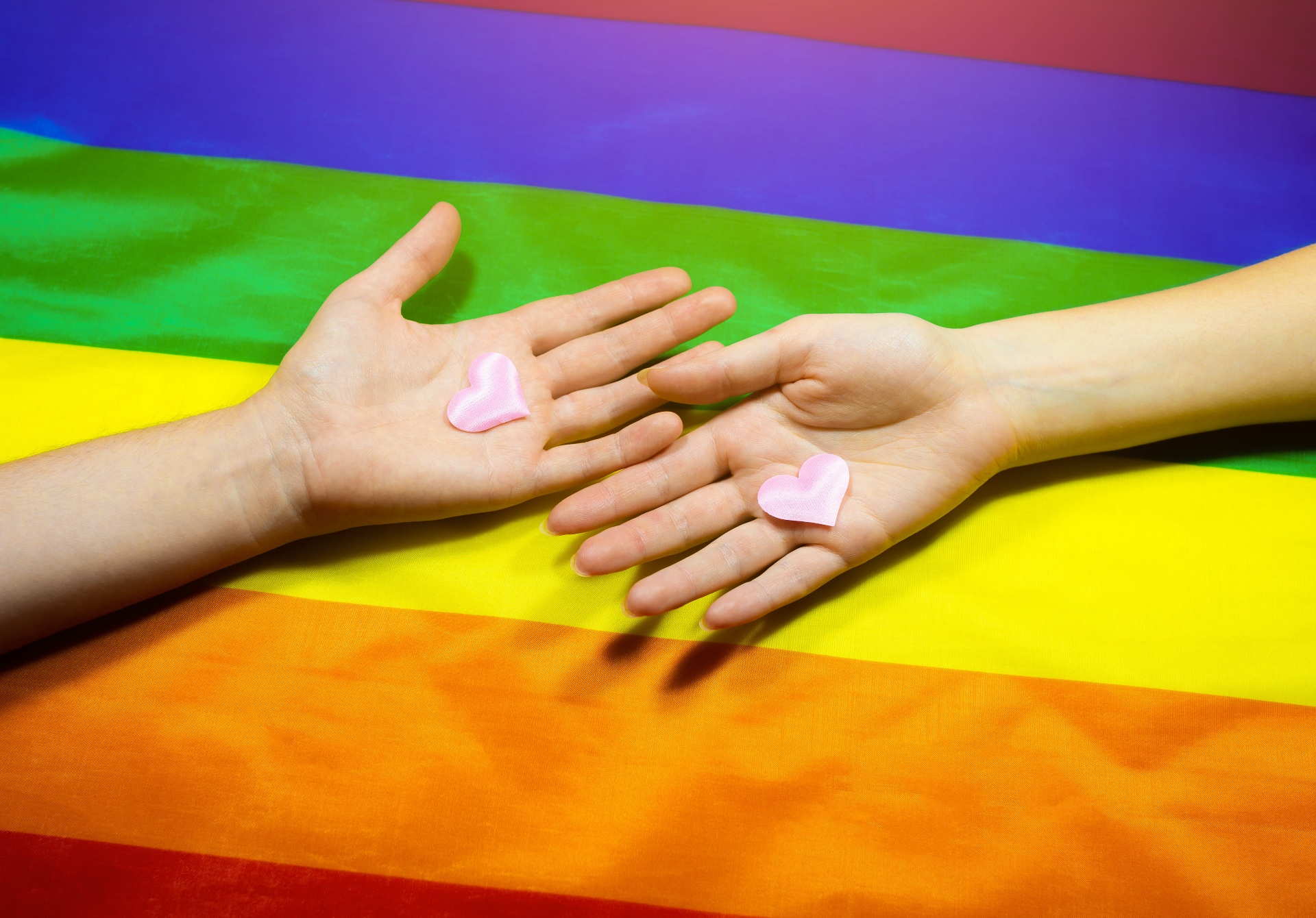 Two hands reaching toward each other with small pink hearts on a rainbow pride flag